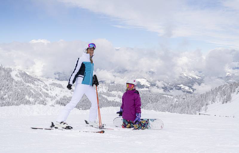 <p>Ein Kind auf einem Snowboard auf Knien vor einer Frau auf Skiern. Die Skipiste ist perfekt präpariert und die Landschaft rundherum tief verschneit. Der Himmel ist blau nur über dem Tal sind ein paar Wolken zu sehen. Dieses Bild entstand im Skigebiet Nassfeld in Kärnten, dem Skigebiet an der Grenze zu Italien, welches auch zu den "TOP 10 Skigebieten" Österreichs gehört.</p>