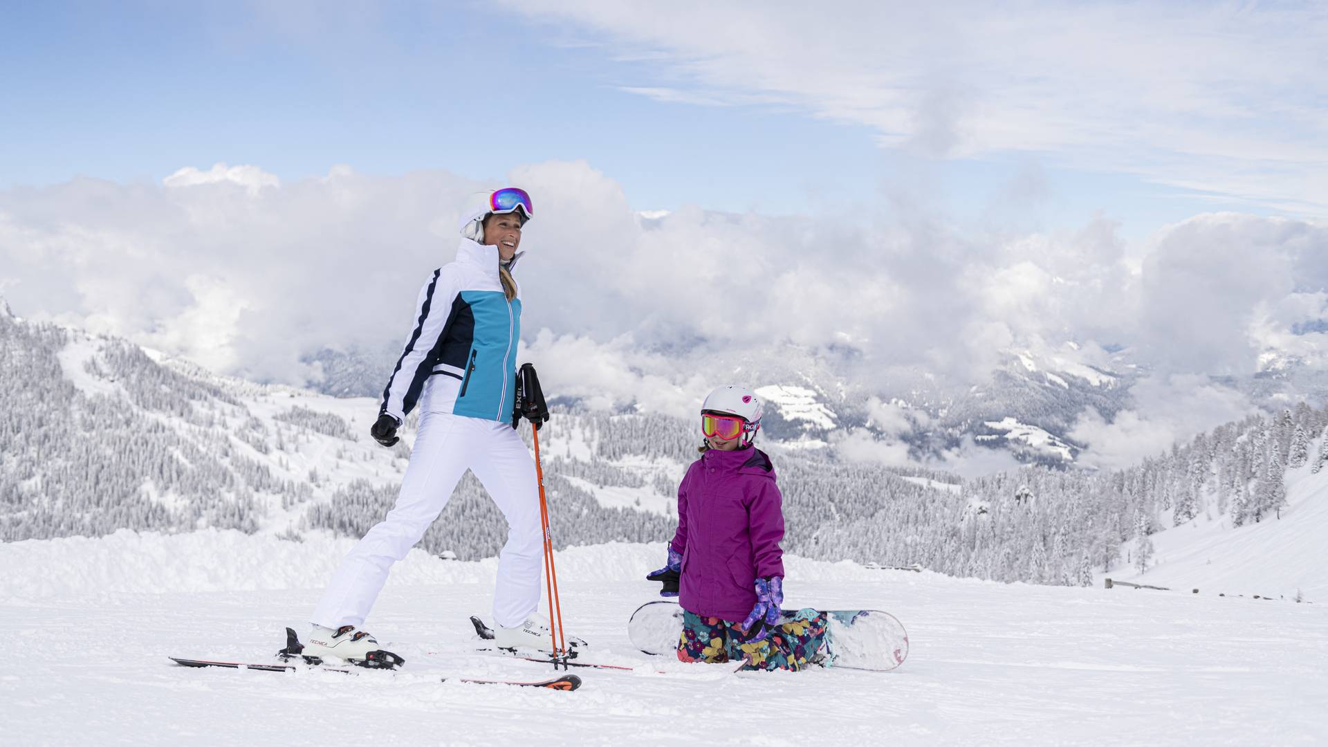 <p>Ein Kind auf einem Snowboard auf Knien vor einer Frau auf Skiern. Die Skipiste ist perfekt präpariert und die Landschaft rundherum tief verschneit. Der Himmel ist blau nur über dem Tal sind ein paar Wolken zu sehen. Dieses Bild entstand im Skigebiet Nassfeld in Kärnten, dem Skigebiet an der Grenze zu Italien, welches auch zu den "TOP 10 Skigebieten" Österreichs gehört.</p>